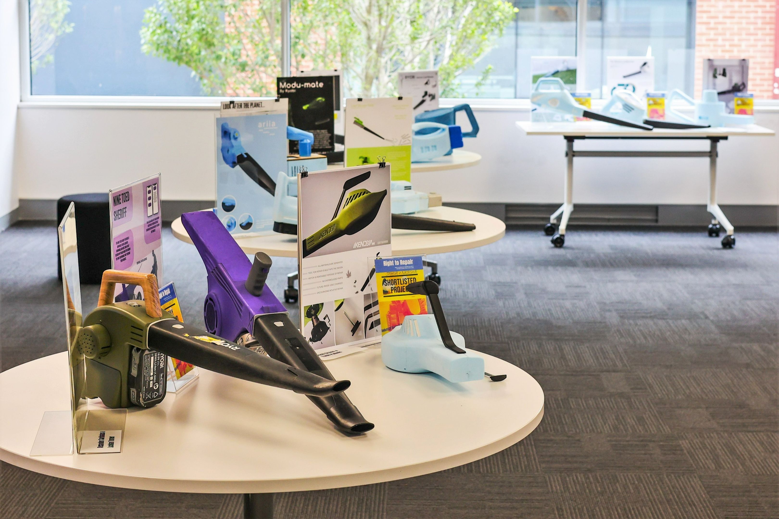 A range of leaf blower prototypes, some made from coloured plastics and others from blue sculpting foam, are displayed on tables next to informative signs