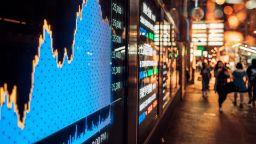 Pedestrians walking outside a shopfront with screens showing stock market activities.