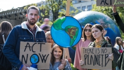 Young people gather outside for a protest on climate action, holding cardboard signage reading 'Act now!' and 'There is no Planet B', and a megaphone.