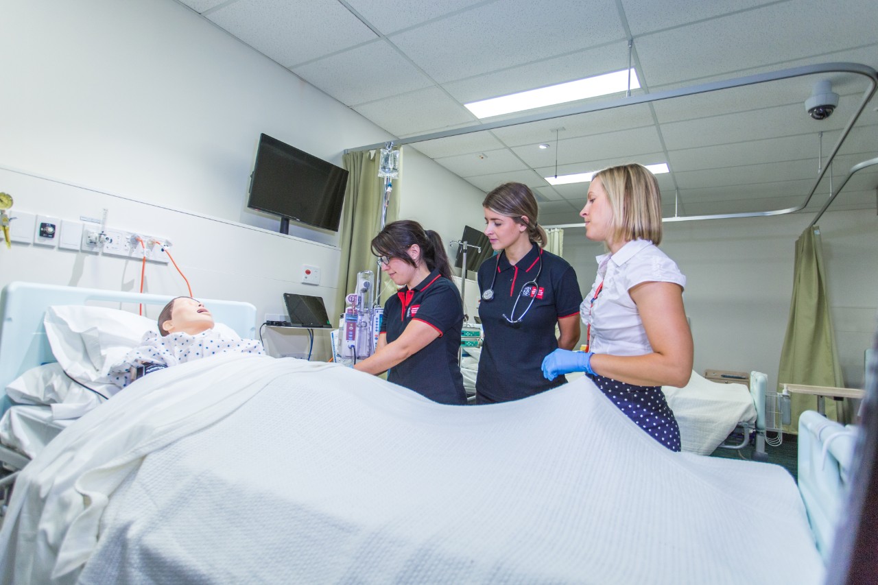 Nursing students at the Hawthorn nursing lab stand by a hospital bed
