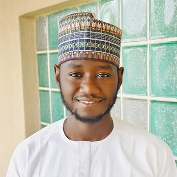 Abubakar in a white shirt and colorful hat standing outside against a beige building.