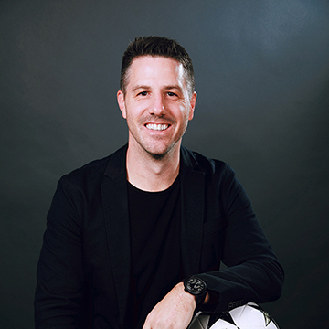 Andrew Collins sitting and smiling in a dark suit against a black background.