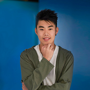 Takumi standing in front of a blue background in a green and grey outfit.