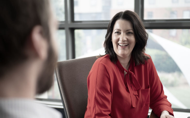 A woman is pictured wearing a red shirt. She is sitting at a desk and speaking to a man who is close up and out of focus. A grey filter has been placed over the image.