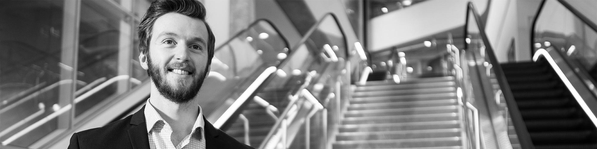 A greyscale image of a man in a suit standing inside a building lobby with escalators behind him.