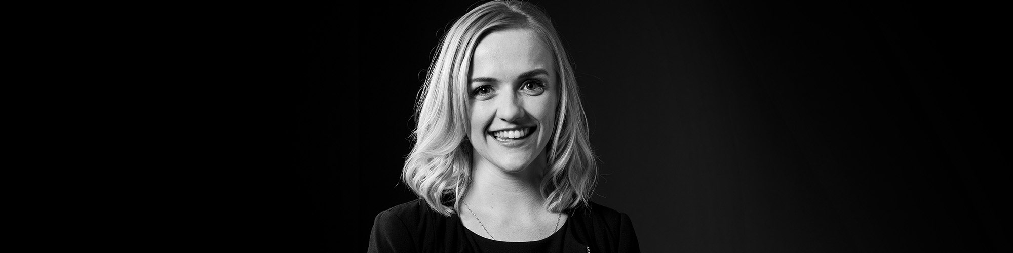A black and white photo of a woman in front of a black background and red square. The woman is smiling and wearing professional attire.