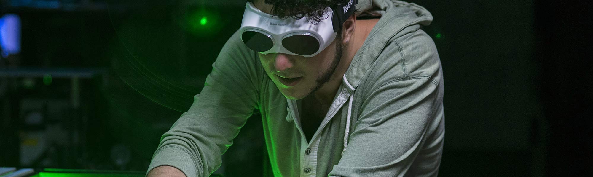 A young man wearing protective eyewear is working on something off camera. Behind him is a dark room and green light.